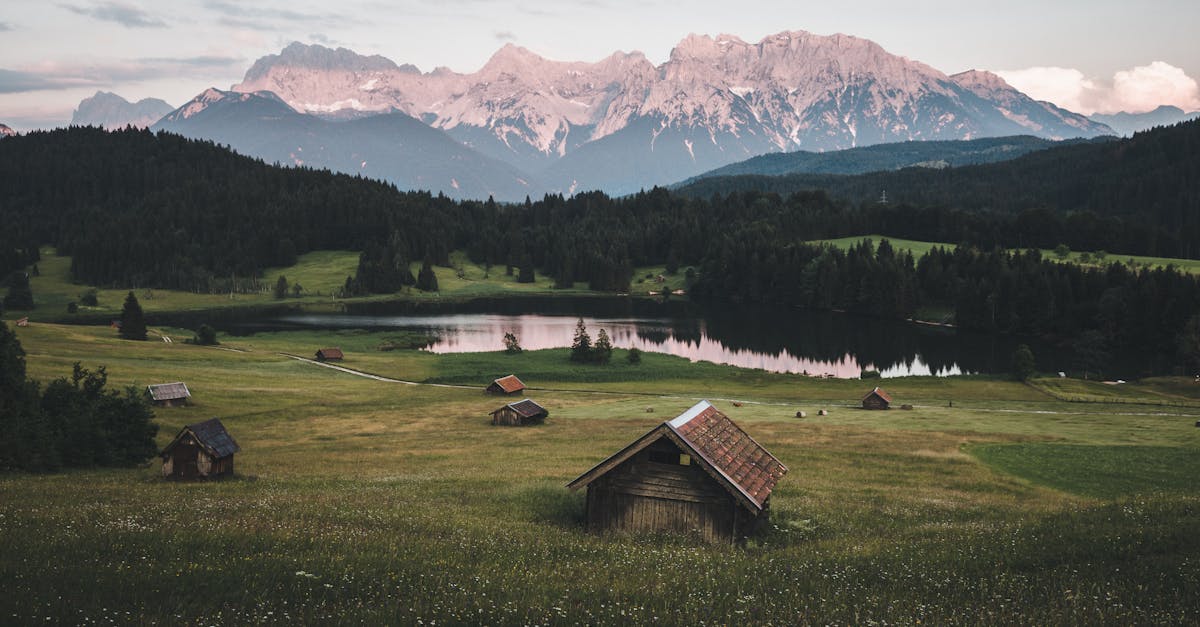 Explore the serene Bavarian landscape in Krün, Germany, with lush fields, a tranquil lake, and majestic mountains at dawn.