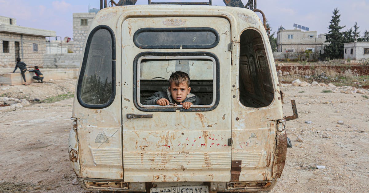 A young boy peers from an abandoned van in the desolate streets of Idlib, Syria, amidst war devastation.